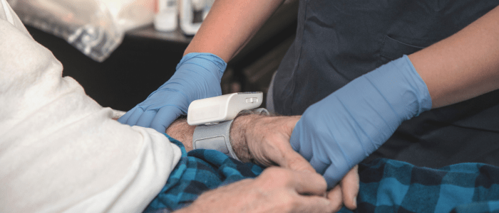An older persons arm with a monitoring device on the wrist, their hand being held by a nurse in blue latex gloves. To show the concept of new guidance on wearable devices to remotely monitor Parkinson's.
