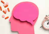 A cut out of a brain made of pink paper on top of a cut out of a head made of pink paper. To the right of the head is an alarm clock, to the left is several pink tablets and red capsules. To represent the concept of erectile dysfunction medications linked to lower Alzheimers risk.