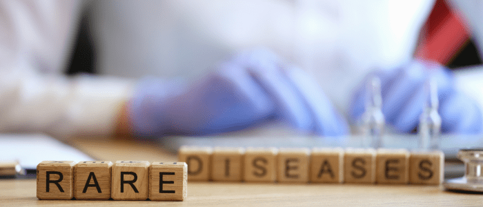 Small wooden blocks spelling out 'rare diseases' in front of a person in a lab coat wearing blue gloves using a test tube. To show the concept of a new report offers insights on care and research for rare diseases.