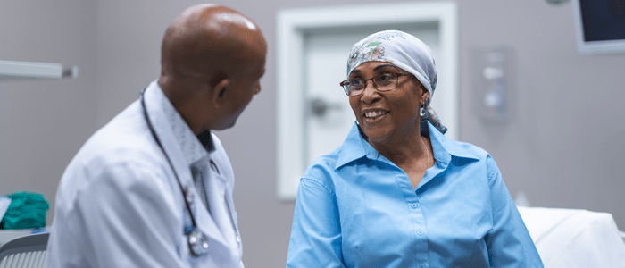 A woman receiving cancer treatment sits on a hospital bed and smiles as a Dr talks to her. To represent that Flatiron Health and Smirta partner to enhance oncology scheduling.