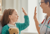 A nurse wearing a light blue uniform and a stethoscope high fives a young girl holding a teddy bear. The girl is sitting on her mothers lap, and they are all smiling. To represent that Truveta releases the largest mother–child EHR dataset.
