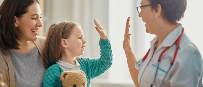 A nurse wearing a light blue uniform and a stethoscope high fives a young girl holding a teddy bear. The girl is sitting on her mothers lap, and they are all smiling. To represent that Truveta releases the largest mother–child EHR dataset.