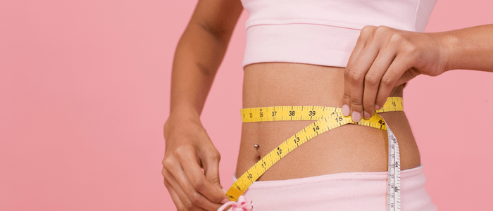 A woman wearing pink clothes measures her waist with a yellow tape measure. The image has a pink background. To represent the concept of GLP-1 RA anti-obesity medication trends in Medicare-aged adults.