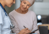 A Dr in a lab coat and wearing a stethoscope writes on a tablet. She is sitting next to a cancer patient with a scarf wrapped around her head. The patient is also looking at the tablet. To represent that Caris and COTA partner for oncology drug development with RWD